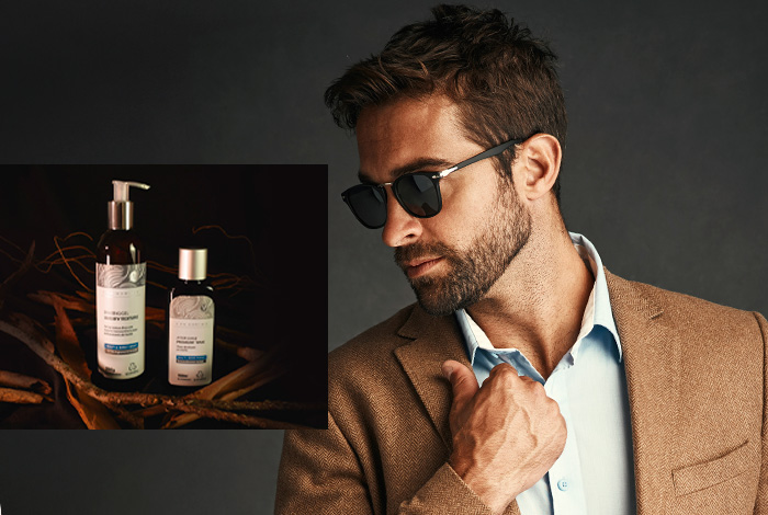 Homem estiloso de barba e óculos escuros ajustando o paletó, com destaque para produtos de cuidados masculinos ao fundo, sugerindo sofisticação.